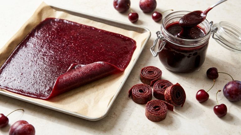 Homemade Persian lavashak: a thin sheet of deep ruby sour-cherry fruit puree dried on a parchment-lined baking sheet with one corner peeled back, beside finished lavashak strips rolled into spirals and an open jar of thick fruit rob with a spoon lifting a ribbon of it